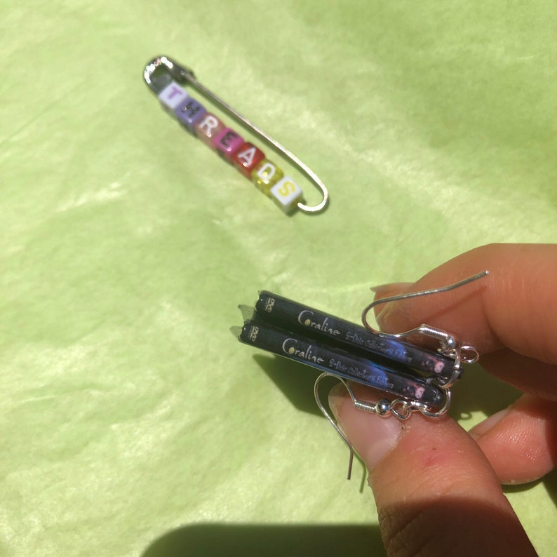 May include: A silver safety pin with the word "THREADS" spelled out in colorful letter beads. Below, a pair of earrings featuring miniature book covers with the title "Coraline" are visible.