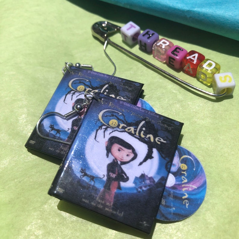 May include: Miniature Coraline DVD earrings and a matching CD charm. The earrings feature the movie cover art with a heart-shaped hook. A safety pin with the word "THREAD" spelled out in colorful beads is also in the image.