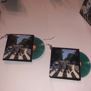 Puede incluir: Un par de pendientes con una imagen cuadrada de la portada del álbum de los Beatles "Abbey Road" con un disco de vinilo verde unido a cada pendiente.