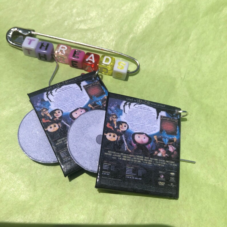 May include: A handmade pin with the word "THREADS" spelled out in colorful letter beads. Attached are miniature DVD cases and silver-colored CDs, featuring a movie poster design. The items rest on a light green surface.
