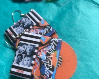 May include: A pair of earrings featuring a black and white striped design with a photo of a band on one side and a colorful abstract design on the other side. The earrings are made of metal and have a silver finish. The earrings are hanging from a silver hook.