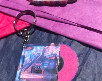 Puede incluir: Un llavero rosa y negro con un disco de vinilo en miniatura y una foto de un edificio con el texto "OCT 18 1985 3:00 PM". El llavero está unido a un llavero plateado. También se muestra un imperdible con la palabra "THREADS" escrita en cuentas de colores.