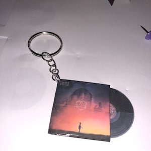 Peut inclure: Un porte-clés argenté avec une miniature de pochette d'album attachée. La pochette d'album est pour l'album "In Return" du groupe "ODESZA". La pochette d'album présente un motif géométrique et une personne debout dans un champ.
