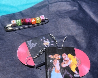 May include: A pair of silver earrings with black and white album covers featuring a band called "The 1975". The album covers are attached to the earrings with silver chains. The earrings are on a black surface with a pink CD case.