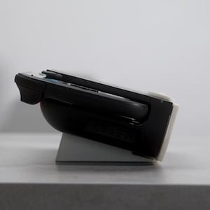 May include: A black handheld gaming device is docked on a white and gray stand. The device is angled, showing its sleek design. The stand provides support and stability for the device.
