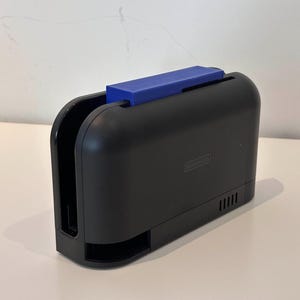 May include: Black Nintendo Switch dock with a blue insert. The dock has a rounded design with the Nintendo logo on the front. The dock is sitting on a white surface.