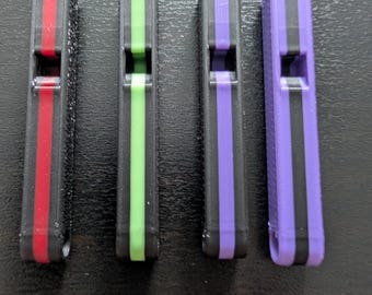 May include: Four rectangular whistles in a row, each with a black base and a colored stripe down the center. The stripes are red, green, purple, and purple. The whistles are made of plastic.