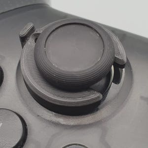 May include: Close-up of a black controller with a textured joystick and surrounding components. The joystick has a circular design with a ridged edge. The controller's surface is a dark gray color.