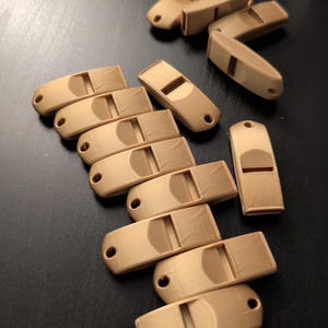 May include: A collection of light brown wooden whistles, each with a rectangular body, a sound hole, and a small hole for a string. The whistles are scattered on a dark surface.