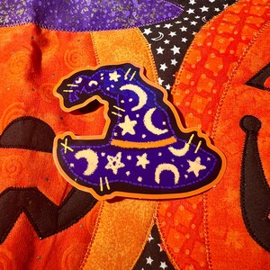 May include: A sticker of a witch hat with a purple crown, decorated with crescent moons and stars. The hat has an orange band and is outlined in black. The sticker is placed on a patchwork background of orange, purple, and black fabrics.