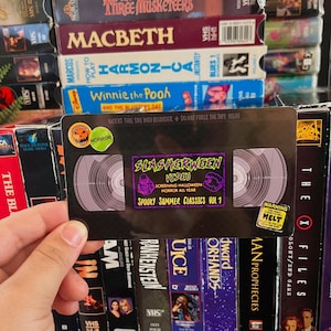 May include: A black rectangular magnet designed to look like a VHS tape. The center label reads "SLASHERWEEN VIDEO SCREENING HALLOWEEN HORROR ALL YEAR SPOOKY SUMMER CLASSICS VOL 1". It features small circular stickers and a yellow warning label.