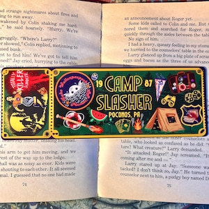 May include: A rectangular metal sign with a green border and the words "Camp Slasher Pocono, PA" in yellow and black. The sign features illustrations of a tent, a watermelon slice, playing cards, and a vintage suitcase. The sign also has the text "Totally Killer Summer".