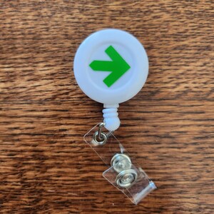 May include: White retractable badge holder with a green arrow pointing to the right. The holder has a clear plastic strap with two metal clips.