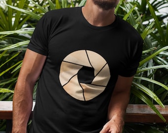 Może przedstawiać: Czarny t-shirt z beżowym nadrukiem obiektywu aparatu. Nadruk jest stylizowanym przedstawieniem obiektywu aparatu z wieloma listwami.