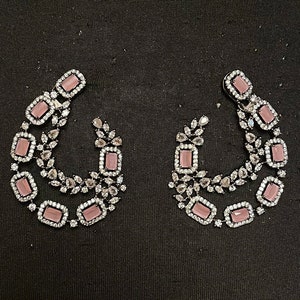 Puede incluir: Un par de pendientes plateados en forma de lágrima. Cada pendiente presenta una serie de piedras preciosas rosas rectangulares rodeadas de piedras claras más pequeñas. El diseño incluye detalles en forma de hojas, creando un aspecto elegante.