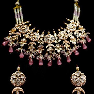 Può includere: Un set di collana e orecchini da sposa indiani in oro e rosa con accenti di perle e pietre rosa. La collana presenta un grande ciondolo decorato con un motivo a mezzaluna. Gli orecchini sono progettati in modo simile con un ciondolo più piccolo e pietre rosa pendenti.