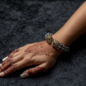 May include: A hand with henna designs and white nail polish is wearing a silver and gold bracelet with a large, round, clear stone in the centre.