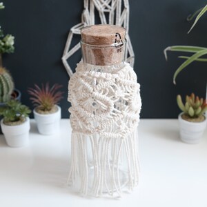 Könnte beinhalten: Ein klares Glas mit Korken, umhüllt von einer cremefarbenen Makramee-Hülle mit Fransen. Das Glas steht auf einer weißen Oberfläche, mit Pflanzen im Hintergrund.