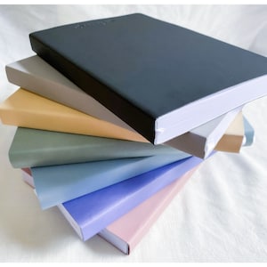 Peut inclure: Une pile de sept cahiers de différentes couleurs, dont noir, gris, beige, vert, bleu, violet et rose. Les cahiers sont tous fermés et empilés les uns sur les autres.