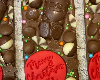 May include: A rectangular milk chocolate bar topped with a moulded chocolate Santa Claus, KitKat pieces, round chocolate spheres, and star-shaped chocolate bits. A red circular disc features the embossed text Merry Christmas MISS GALVIN.