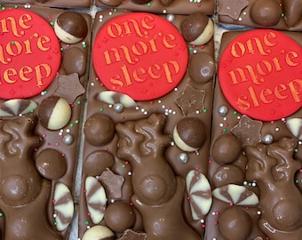 May include: Milk chocolate bars decorated with a red circular disc embossed with the text one more sleep, a moulded chocolate reindeer, round chocolate spheres, star-shaped chocolate pieces, and small multi-coloured sugar sprinkles.