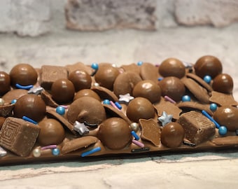 May include: A chocolate bar with a variety of toppings, including chocolate balls, sprinkles, and stars. The bar is a light brown color and has a smooth, glossy finish.