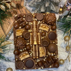 May include: A rectangular milk chocolate bar topped with a central gold-coloured nutcracker figure, surrounded by various chocolate pieces including hazelnut truffles, caramel-filled wafers, and small chocolate spheres, all sprinkled with chopped nuts.