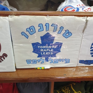 May include: A white box with a blue maple leaf logo and the text "Toronto Maple Leafs" in English and Hebrew. The box is on a wooden shelf.