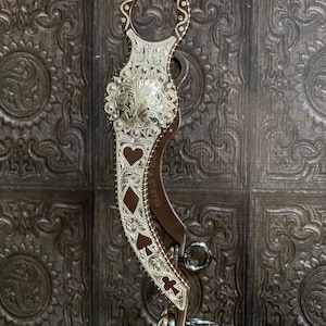 Könnte beinhalten: Ein silber-braunes Pferdgebiss mit aufwendigen floralen Mustern und Spielkartensymbolen. Das Gebiss hat eine dekorative Silberplatte mit Herz-, Karo-, Pik- und Kreuz-Designs. Hergestellt aus Metall und Leder.