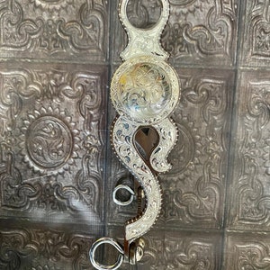 Puede incluir: Un bocado de caballo plateado con intrincados grabados florales y de volutas. El bocado presenta una placa central redondeada y piezas laterales curvas, con anillas para las riendas. La metalurgia es muy detallada, mostrando un tema ecuestre occidental.