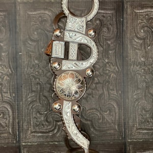 Puede incluir: Un bocado de caballo plateado y marrón con intrincados diseños florales. El bocado tiene una boquilla curva y un anillo en la parte superior.