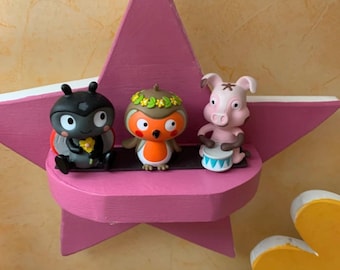 May include: A pink star-shaped shelf displays three cartoon figurines: a black beetle, an orange owl with a green wreath, and a pink pig playing a drum. The shelf is mounted on a wall with a yellow and beige pattern.