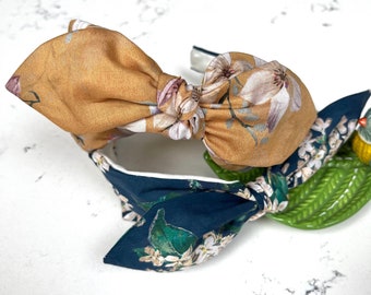 May include: A headband with two large bows, one in a mustard yellow floral print and the other in a dark blue floral print. The headband is made of fabric and has a white elastic band.