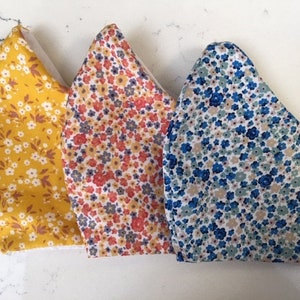 Puede incluir: Tres mascarillas de tela con estampados florales. Las mascarillas son amarillas con flores blancas, naranjas con flores rojas y azules con flores blancas.