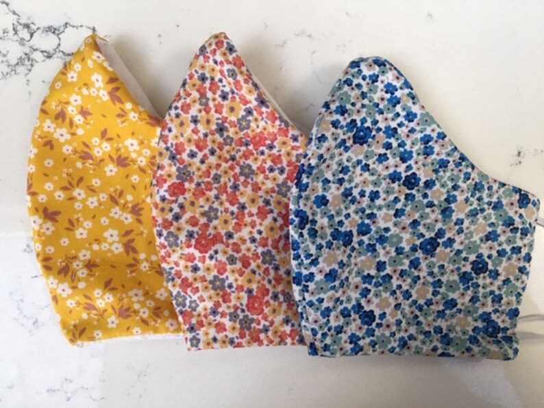 Puede incluir: Tres mascarillas de tela con estampados florales. Las mascarillas son amarillas con flores blancas, naranjas con flores rojas y azules con flores blancas.
