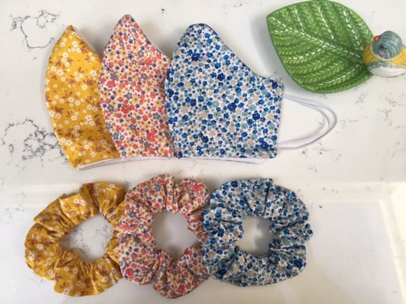 Puede incluir: Tres mascarillas de tela con estampados florales en amarillo, naranja y azul, y tres scrunchies a juego.