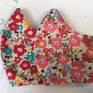 May include: Two face masks with a floral print. The masks are made of a white fabric with red, pink, blue, and yellow flowers.