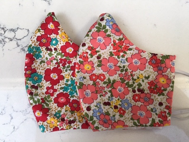 May include: Two face masks with a floral print. The masks are made of a white fabric with red, pink, blue, and yellow flowers.