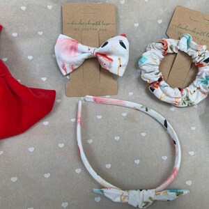 May include: A set of hair accessories featuring a red velvet bow, a white headband with a floral print and a bow, and a white scrunchie with a floral print. The text on the packaging reads "handcrafted with love by PRETTY FAB".