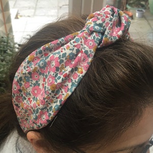 May include: A floral patterned headband with a knotted design. The headband is made of a soft fabric with pink, blue, and yellow flowers on a white background.