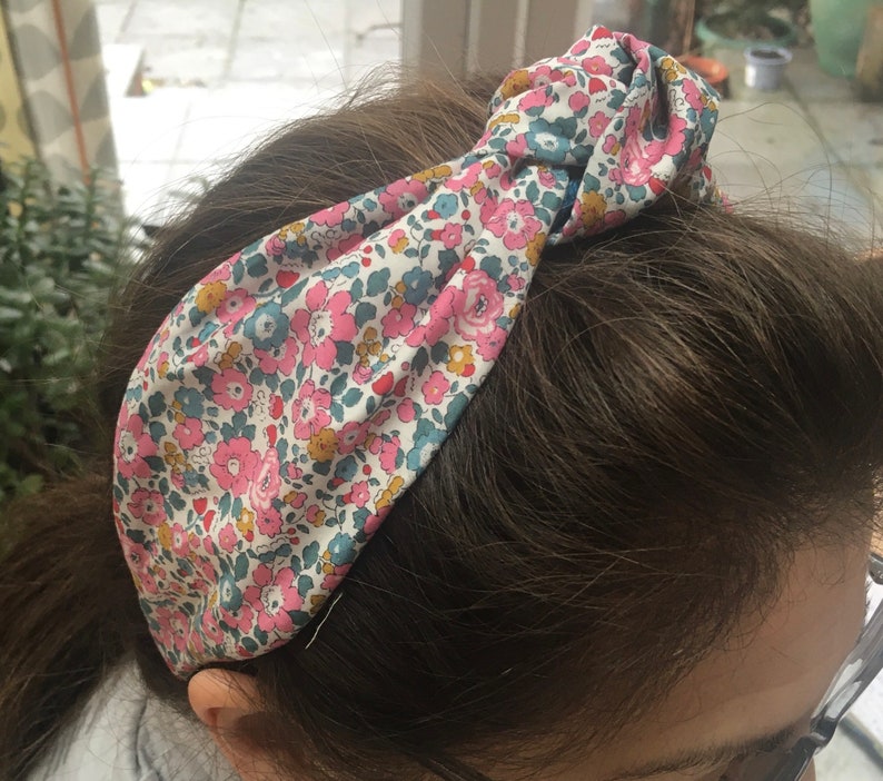 May include: A floral patterned headband with a knotted design. The headband is made of a soft fabric with pink, blue, and yellow flowers on a white background.