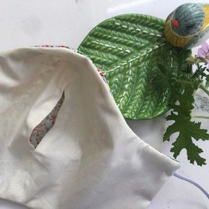 Puede incluir: Una mascarilla facial de algod&oacute;n blanco con un forro de estampado floral. La mascarilla tiene una peque&ntilde;a abertura para la nariz y est&aacute; doblada por la mitad.