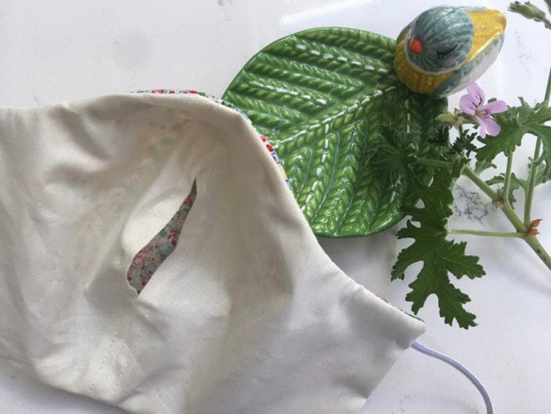 Puede incluir: Una mascarilla facial de algod&oacute;n blanco con un forro de estampado floral. La mascarilla tiene una peque&ntilde;a abertura para la nariz y est&aacute; doblada por la mitad.
