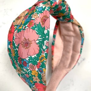 May include: A floral patterned headband with a knotted design. The headband is made of a soft fabric with a pink background and features a variety of colorful flowers.