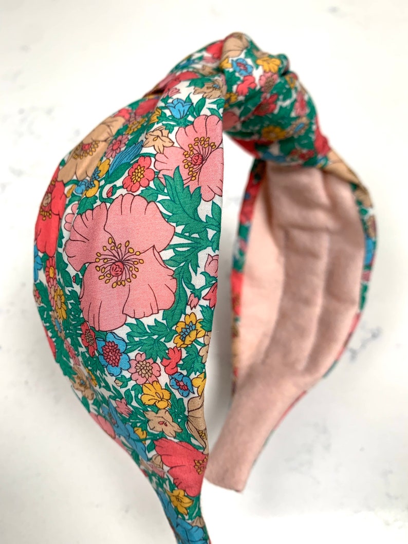 May include: A floral patterned headband with a knotted design. The headband is made of a soft fabric with a pink background and features a variety of colorful flowers.