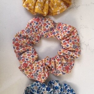 Puede incluir: Tres scrunchies de tela con estampados florales. El scrunchie superior es amarillo con flores blancas, el scrunchie del medio es naranja con flores blancas y azules, y el scrunchie inferior es azul con flores blancas y verdes.