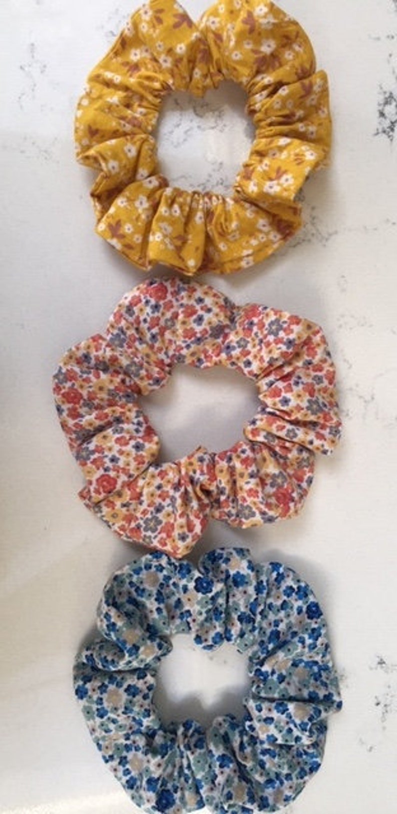 Puede incluir: Tres scrunchies de tela con estampados florales. El scrunchie superior es amarillo con flores blancas, el scrunchie del medio es naranja con flores blancas y azules, y el scrunchie inferior es azul con flores blancas y verdes.