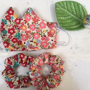 May include: Two floral print scrunchies, one red and one cream, and a floral print face mask with a white background. A green leaf-shaped dish with a bird figurine is also in the image.