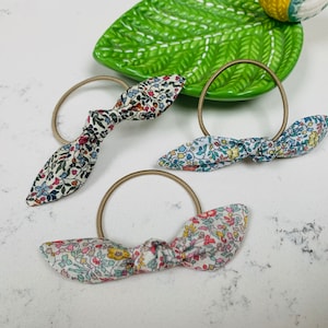 May include: Three hair ties with floral fabric bows. The bows are tied in a knot and the fabric is a white background with small pink, blue, and yellow flowers.