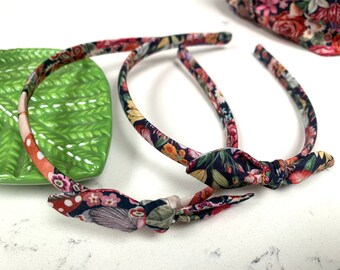 May include: Two floral print headbands with a knotted bow detail. The headbands are made of a dark blue fabric with a colorful floral pattern.